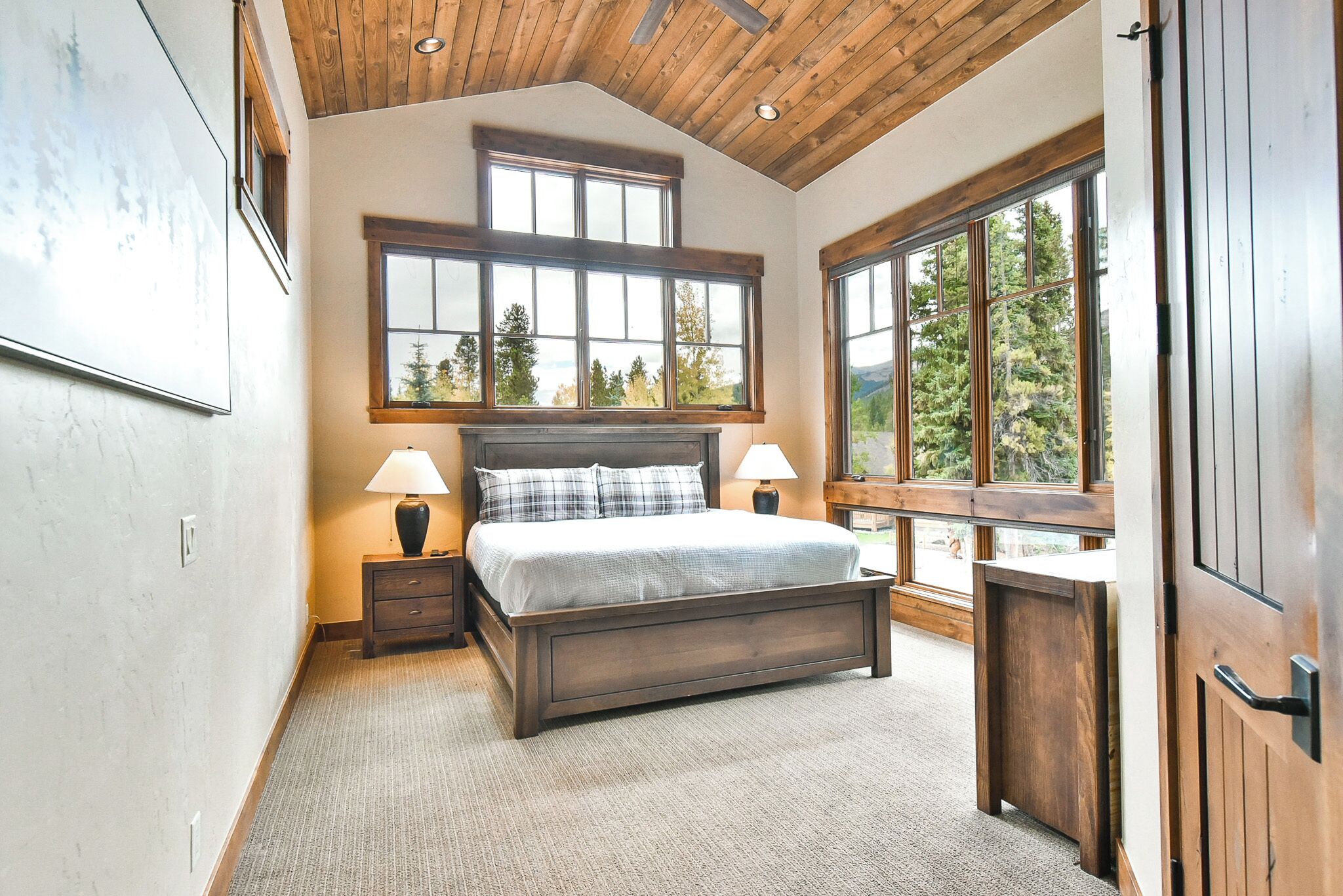 A bedroom with a large bed, two bedside tables with lamps, a wooden headboard, and large windows offering a view of trees.