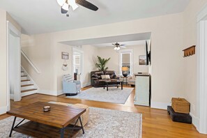 A spacious living room with a wooden coffee table, a sofa, and a dining table. There is a staircase leading to another level, and a kitchen area is visible in the background.