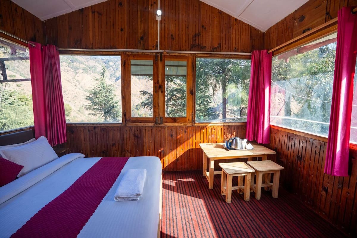 A wooden cabin room with a bed, a table, and a view of trees through large windows.