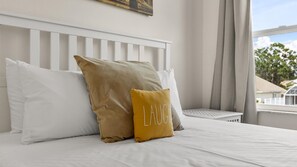 A neatly made bed with white and mustard pillows, a white headboard, and a window with a view of houses and trees. - Upstay: Bright Family-Friendly Retreat with Patio (Kissimmee)