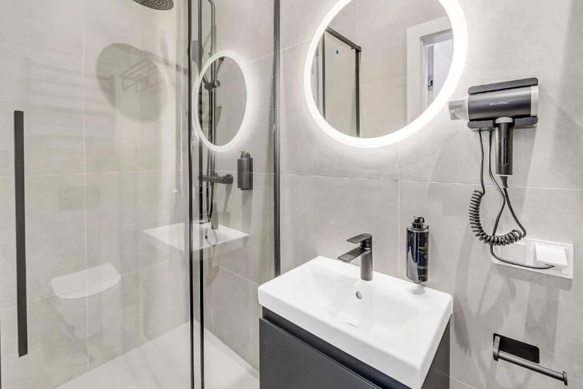 Un baño moderno con cabina de ducha de vidrio, un espejo redondo con iluminación integrada, un lavabo blanco con grifo cromado y un secador de pelo montado en la pared.