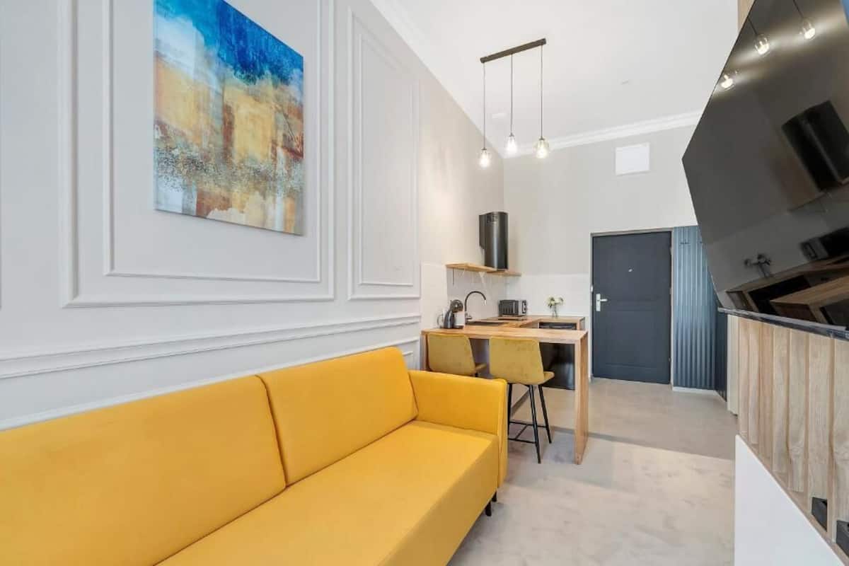 Una sala de estar moderna con un sofá amarillo, un cuadro en la pared y una zona de cocina con estufa y lavaplatos.