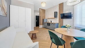Un soggiorno moderno con un divano bianco, un tavolo da pranzo rotondo in legno con sedie verdi, una zona cucina con una TV e un'ampia finestra.