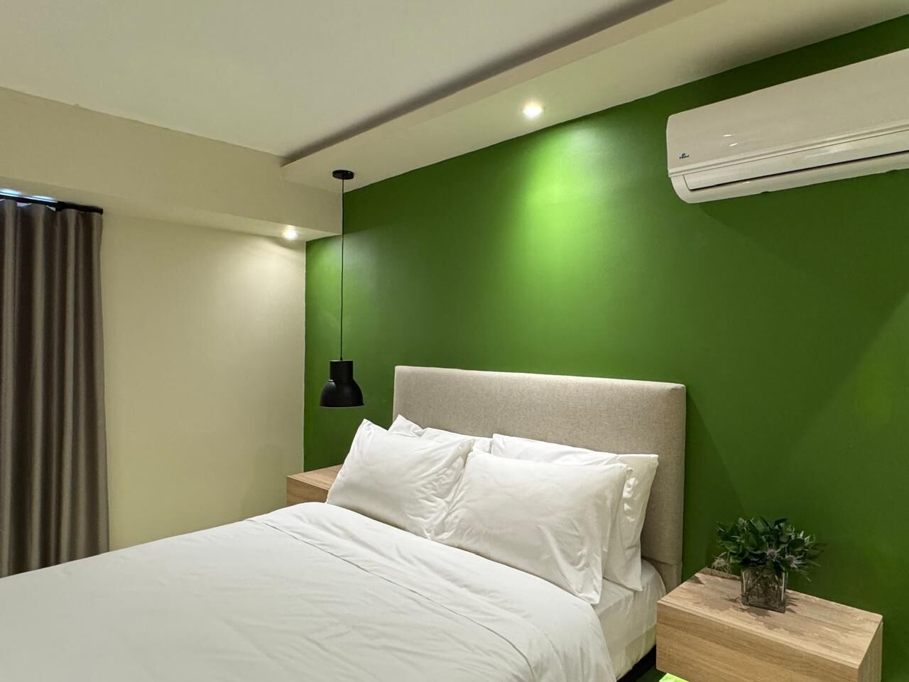 A modern bedroom with a green accent wall, a white bed, a black pendant light, and a small plant on the nightstand.