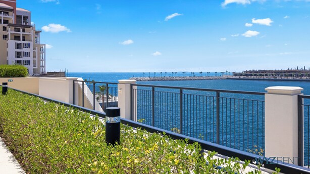 Vista costera con balcón sobre el mar, un paseo marítimo y un paisaje urbano lejano.