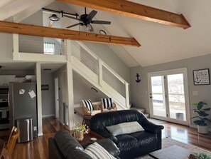 A spacious living area with a high ceiling, wooden beams, a black leather sofa, a dining table, and a view through a glass door.