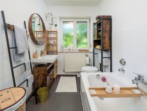 Ein modernes Badezimmer mit einem Holzbadschränkchen, einer freistehenden Badewanne, einer Toilette und einem Fenster mit Blick auf Grünflächen.