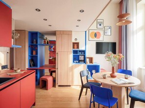 A modern kitchen with a red countertop, blue shelving, and a dining area with a round table and blue chairs. - TurnKey | Vinohrady Modern 2BR Ap. with Balcony (Prague 3)