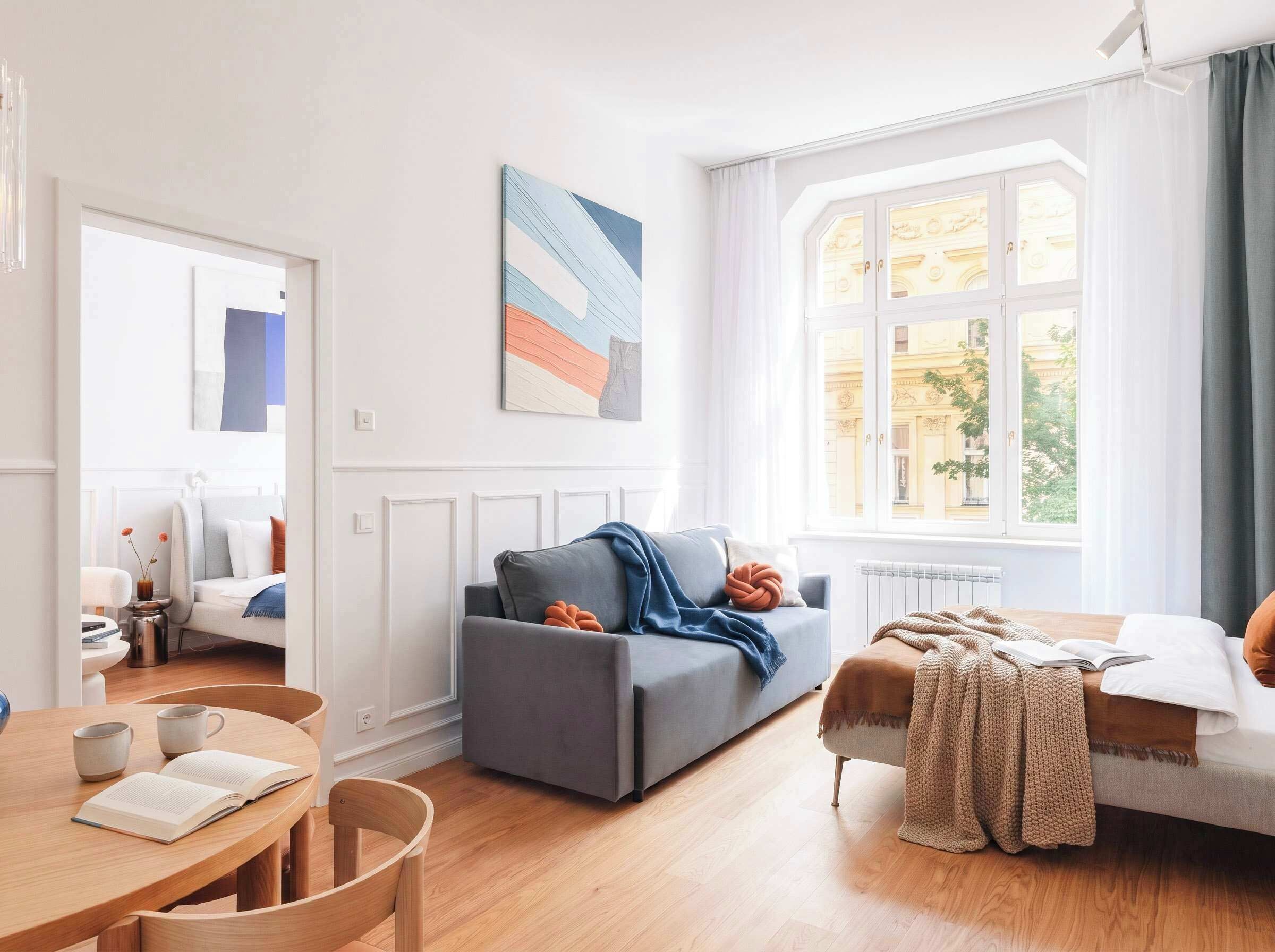 Una sala de estar moderna con un sofá gris, una mesa de centro de madera con libros y un ventanal con vistas a edificios.