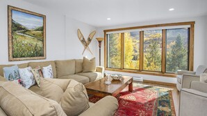 Ein Wohnzimmer mit einem beigefarbenen Sofa, einem Couchtisch und einem großen Fenster mit Blick auf Herbstbäume.