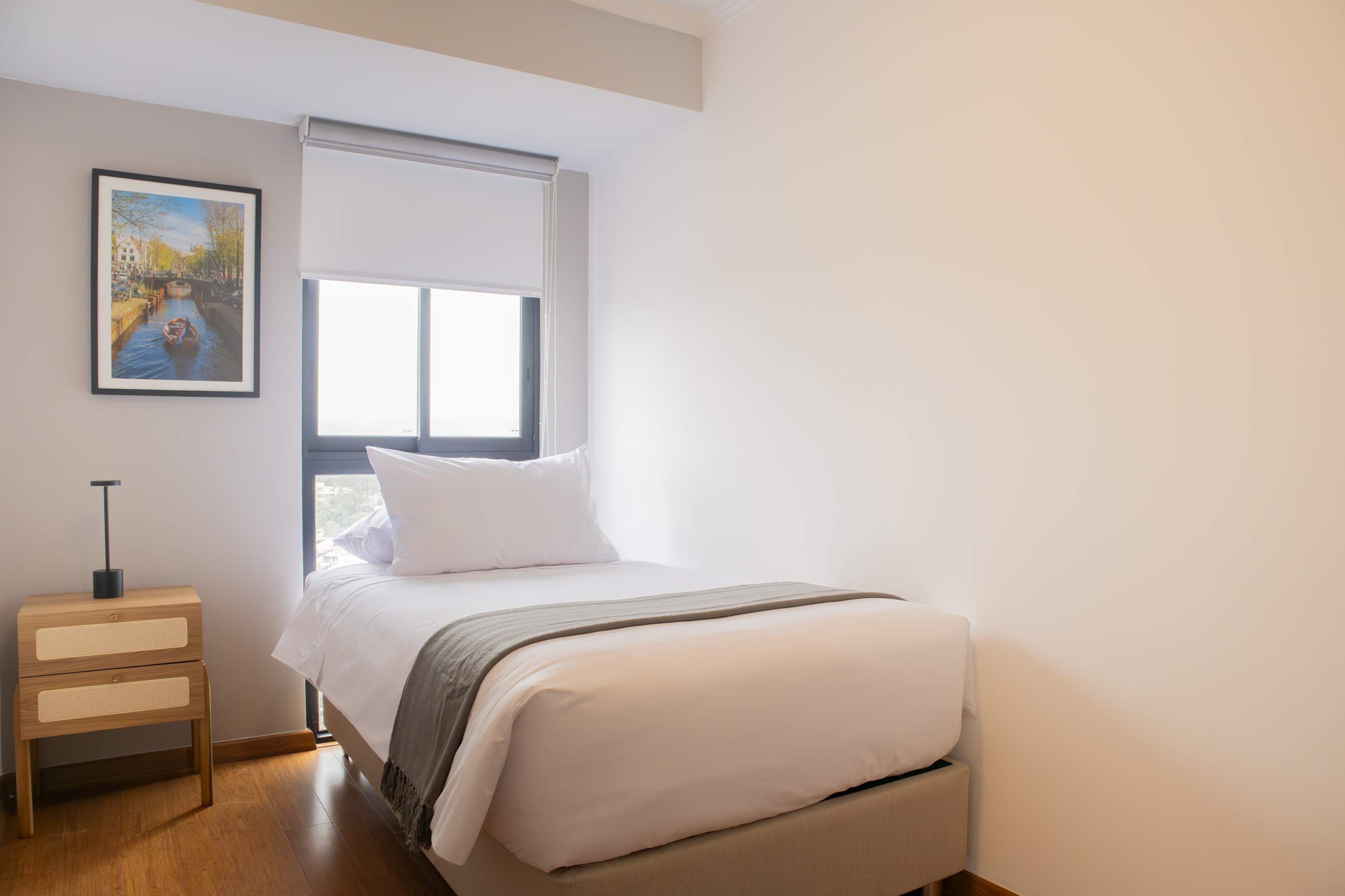 A bedroom with a bed, a nightstand, a window, and a painting on the wall.