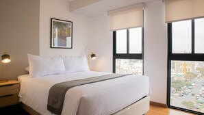 A hotel room with a large bed, a nightstand, a lamp, a framed picture on the wall, and a view of the city through the windows. - w Inviting 2BR w Balcony in Miraflores (Lima)