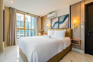 A hotel room with a large bed, a view of buildings through the window, an air conditioning unit, and a bedside table with a lamp.