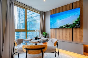 A dining area with a large TV, a table set for two, and a view of a cityscape through a large window.