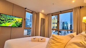 Habitación de hotel con una cama grande, un televisor de pantalla plana en la pared y vistas a la ciudad a través de ventanas amplias.