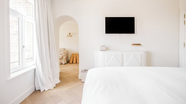 Un dormitorio moderno con una cama grande, un televisor de pantalla plana montado en la pared y una ventana con cortinas blancas.