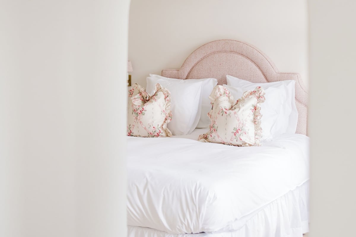 A bedroom with a white bed, pink headboard, and floral pillows.