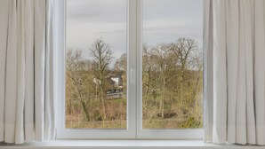 Una ventana con cortinas blancas, que muestra un paisaje rural con árboles y un edificio a lo lejos.
