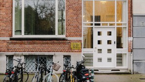 Un edificio de ladrillo con un gran ventanal y una puerta, bicicletas estacionadas al frente y un topogallo verde en la acera.