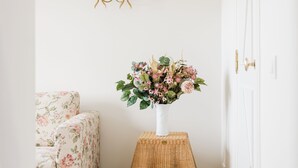 Un pasillo con una puerta blanca, un sillón floral, una mesita con un jarrón de flores y una lámpara fijada a la pared.