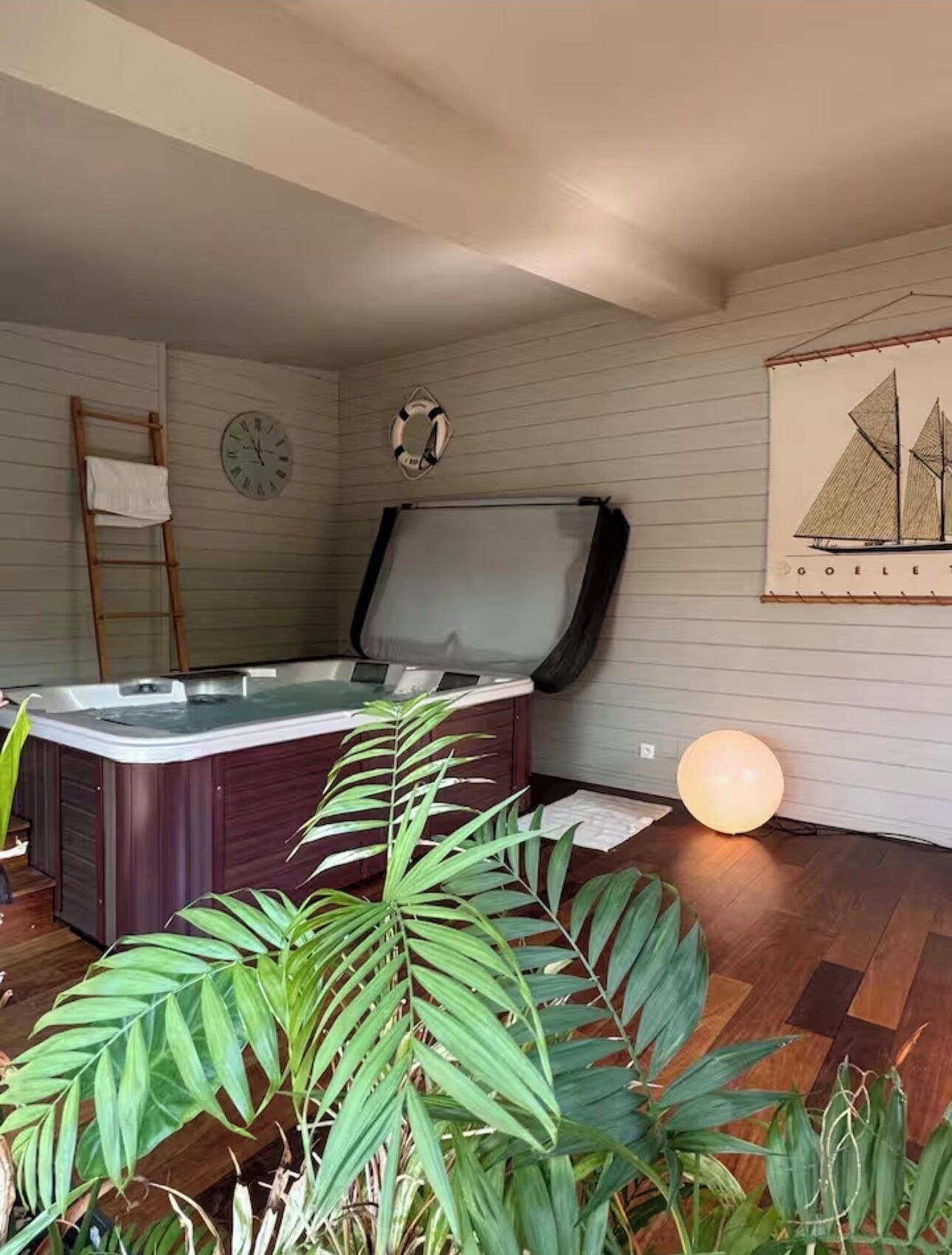 A room with a hot tub, wooden flooring, a ladder, a clock, a life preserver, and a sailboat painting.