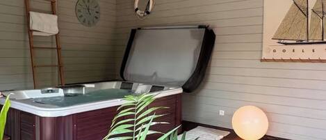 A room with a hot tub, wooden flooring, a ladder, a clock, a life preserver, and a sailboat painting.