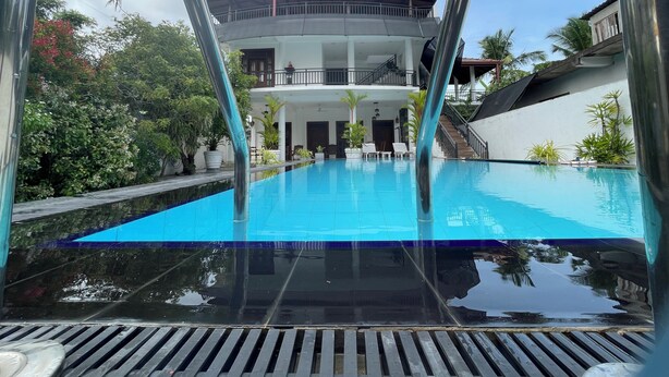 Una piscina con superficie de agua azul cristalina, rodeada de plantas en macetas y un edificio de dos pisos con balcones.