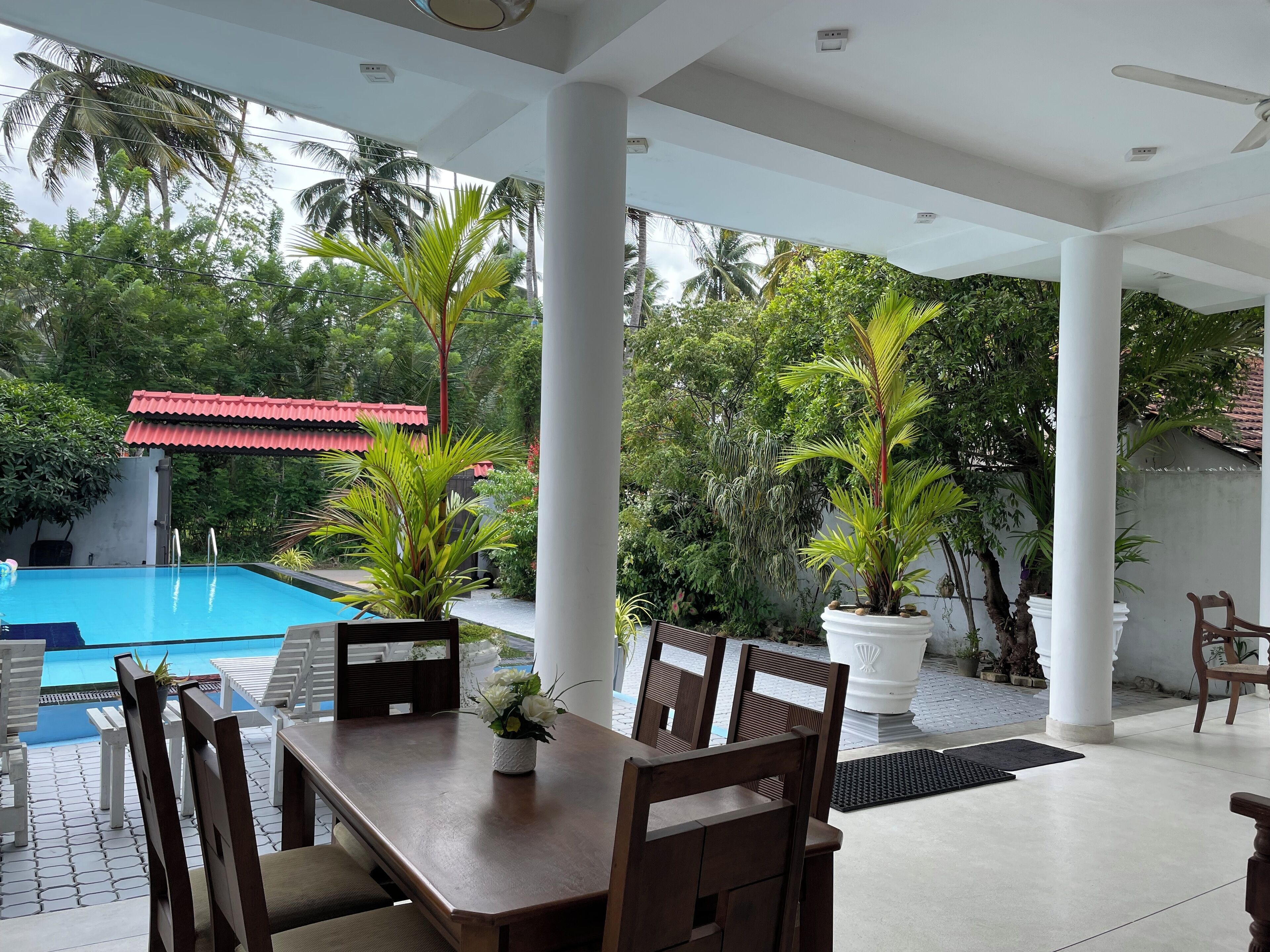 Un área junto a la piscina con mesa y sillas de madera, rodeada de columnas blancas y plantas en macetas.