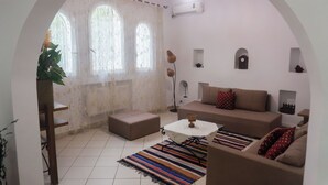 A living room with a sofa, ottoman, coffee table, and a striped rug. There are three windows with curtains, a plant, and a clock on the wall.