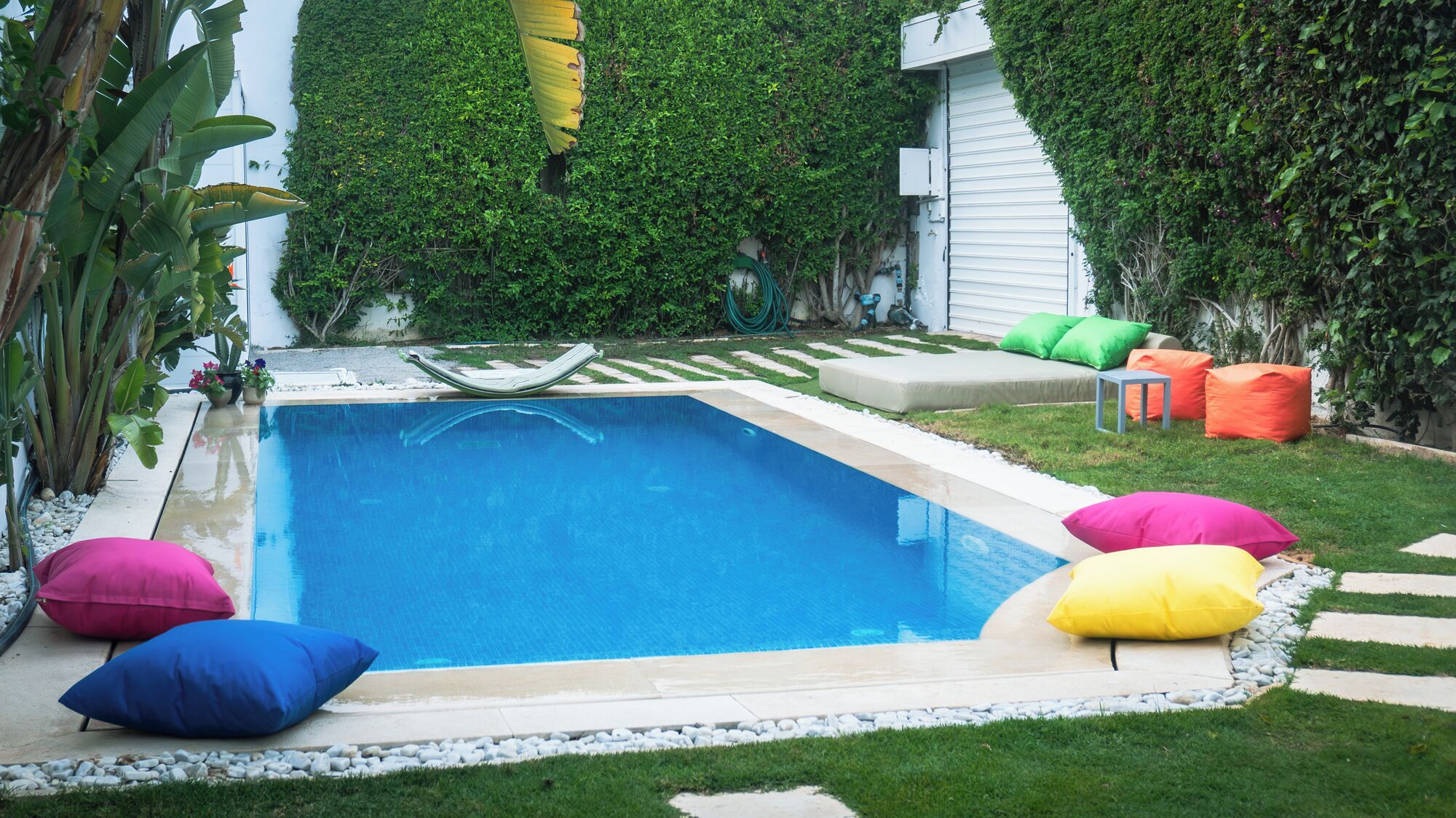 Une piscine rectangulaire entourée de coussins colorés, avec une pelouse et un bâtiment blanc en arrière-plan.