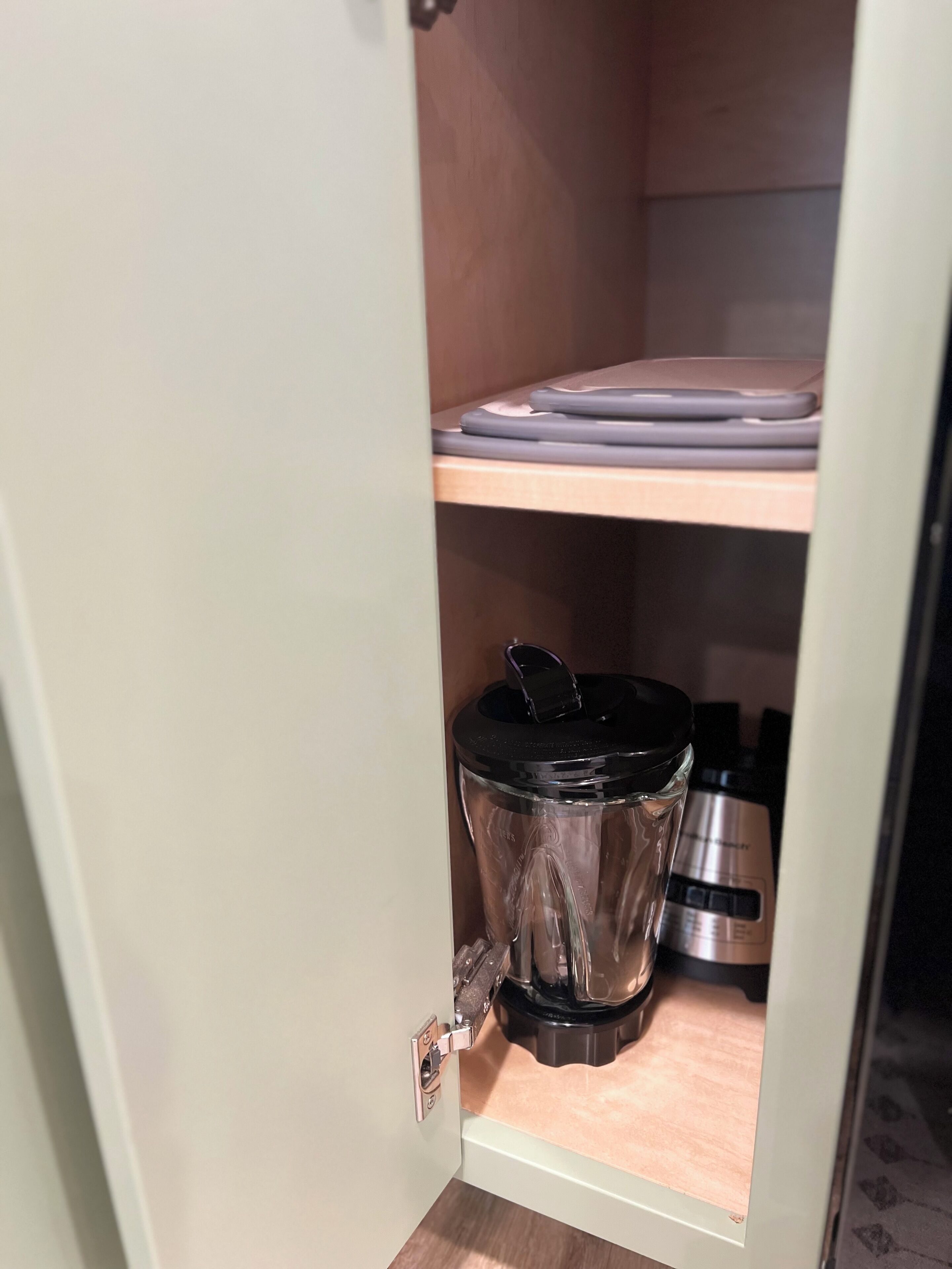 A kitchen with a blender on the counter, a cutting board on the shelf, and a stainless steel appliance below.