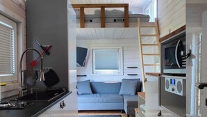 Une cuisine compacte équipée d’une cuisinière, d’un évier et d’un four à micro-ondes. On y trouve un canapé gris avec des coussins, et une fenêtre au-dessus. Une échelle en bois est visible et mène à une mezzanine.