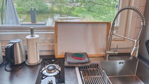 Une cuisine équipée d’une cuisinière à gaz, d’une bouilloire et d’un évier, une fenêtre donnant sur un espace verdoyant et un porte-serviettes.