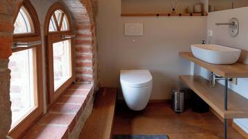 Un baño con inodoro blanco, tocador de madera con lavamanos sobre encimera y ventanal con arco de ladrillo.