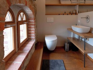Et badeværelse med et hvidt toilet, en trækommode med en skålhåndvask og et vindue med en muråbning.