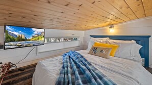 A bedroom with a wooden ceiling, a bed with white bedding and a blue and yellow pillow, a mounted TV displaying a mountain landscape, and a window with blinds.