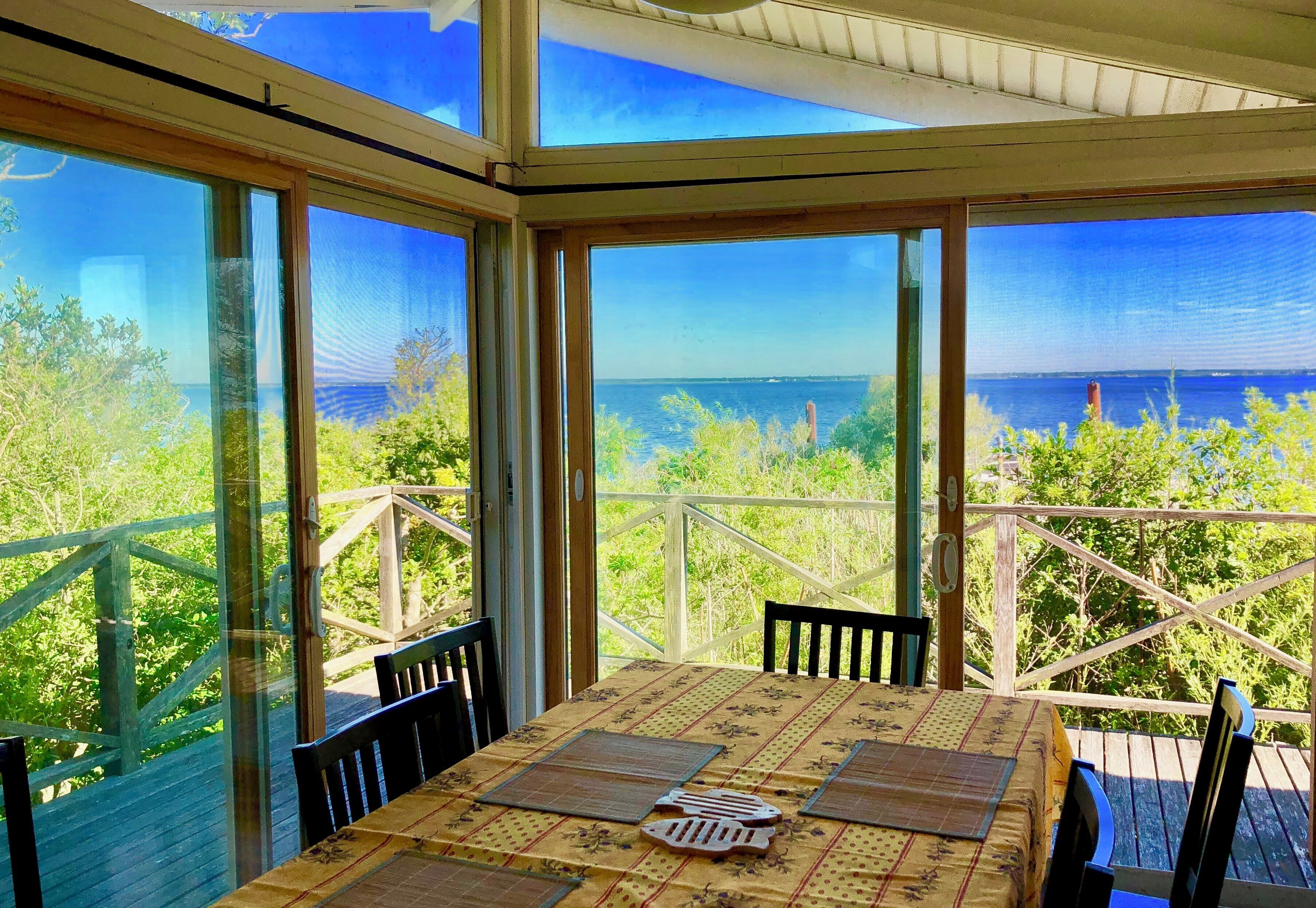 A sunlit room with a table and chairs, large windows offering a view of the sea and greenery.