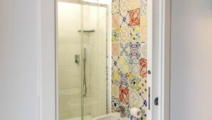 Une salle de bain avec une douche à parois de verre, un lavabo blanc et des toilettes. Les murs sont recouverts de carreaux aux motifs variés.