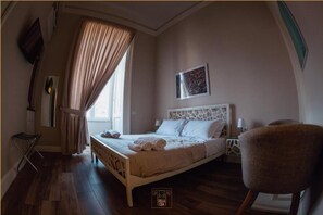 Un dormitorio con cama, una silla, una ventana con cortinas, un cuadro en la pared y un logotipo en la cama.