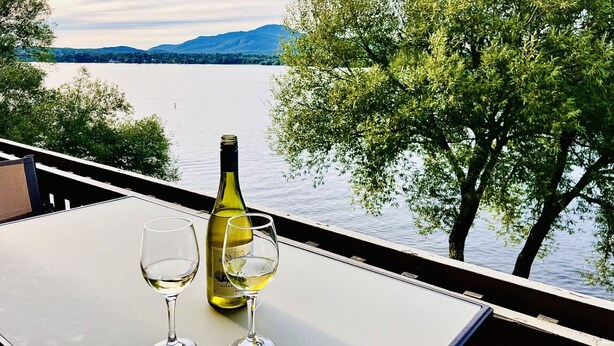 Een tafel met een fles witte wijn en twee glazen, met uitzicht op een meer met bomen en bergen op de achtergrond.