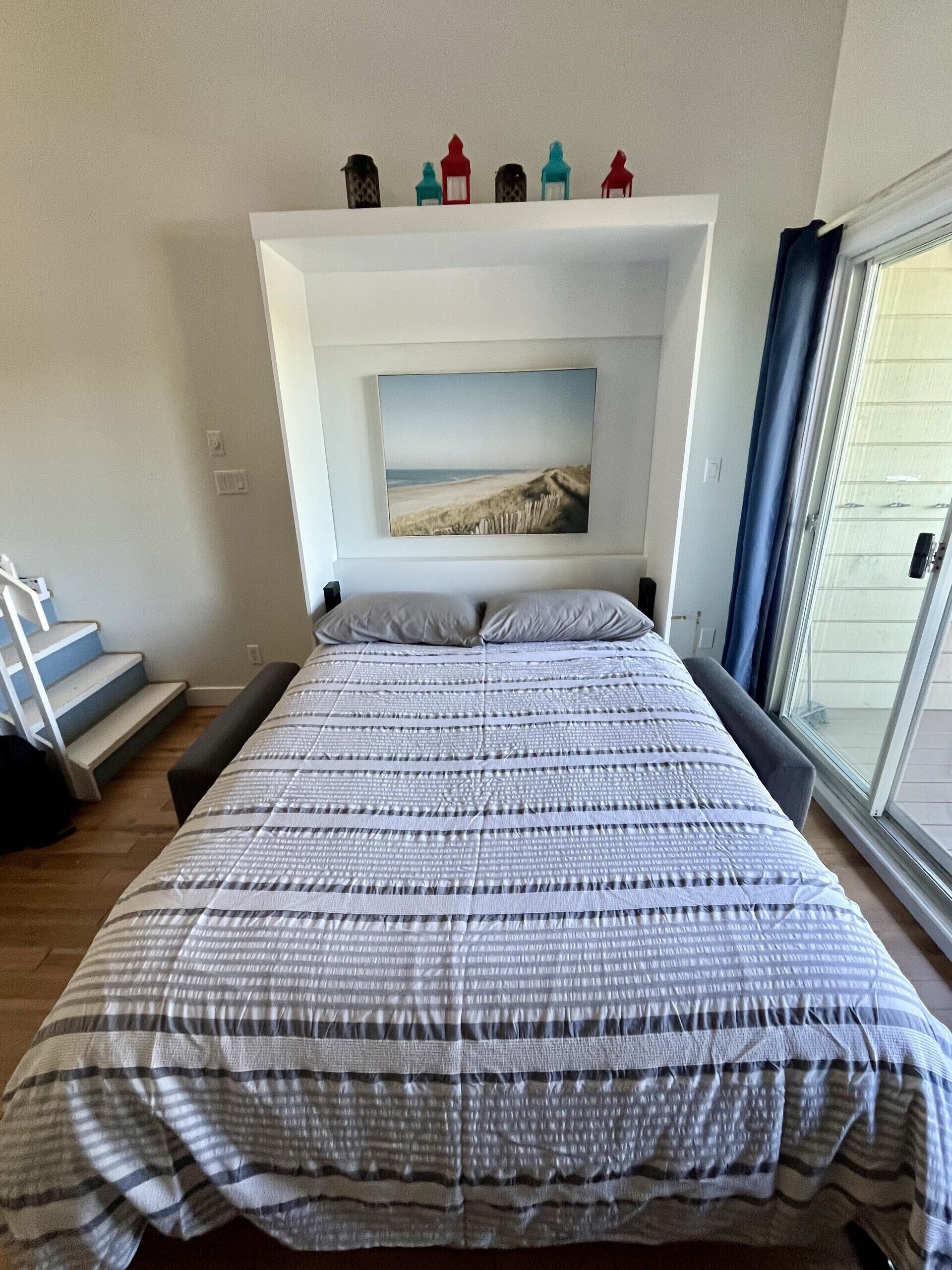 Un dormitori amb un llit, un quadre d'una platja, una escala i una finestra amb cortines.