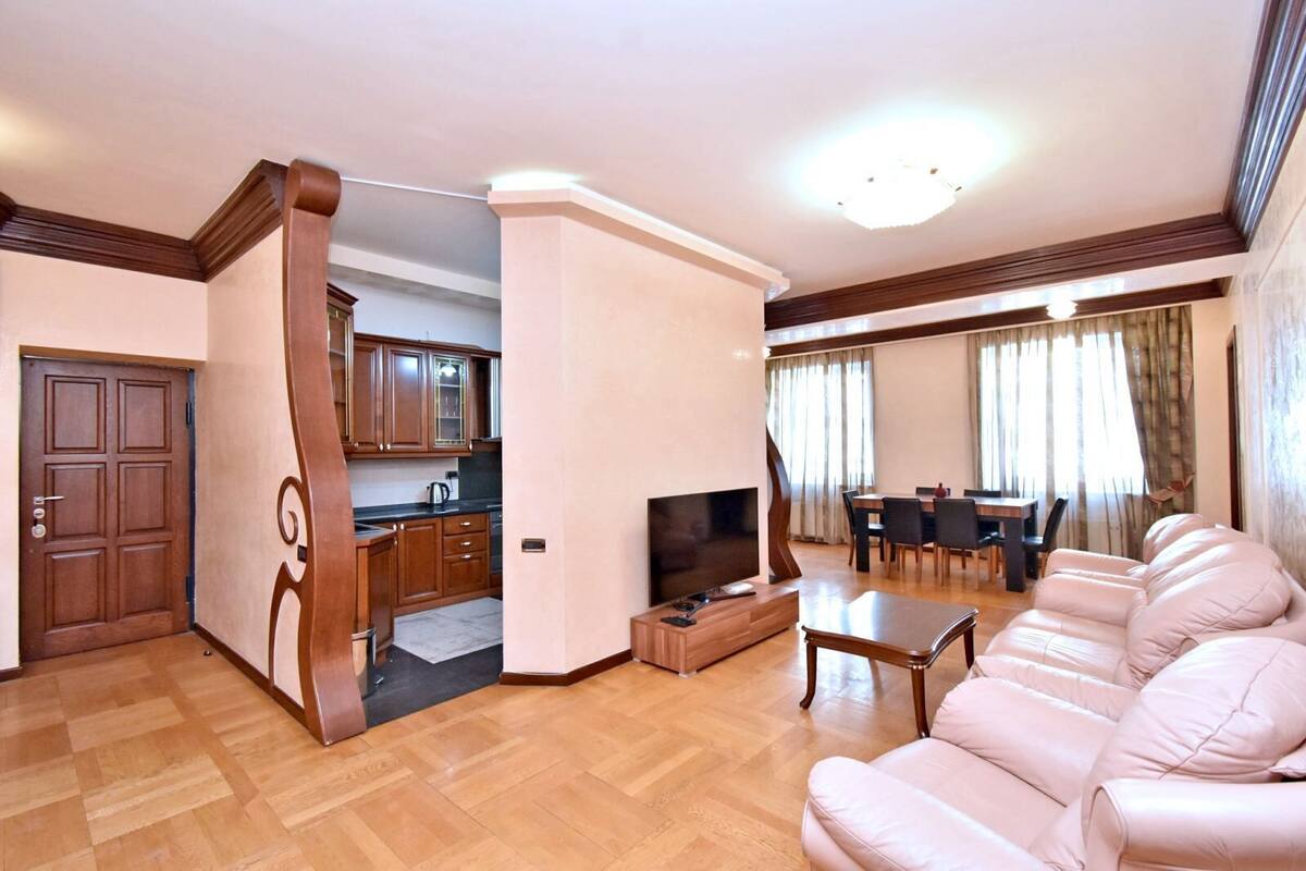 A spacious living room with a large leather sofa, a dining area with a table and chairs, a kitchen with wooden cabinets, and a TV.