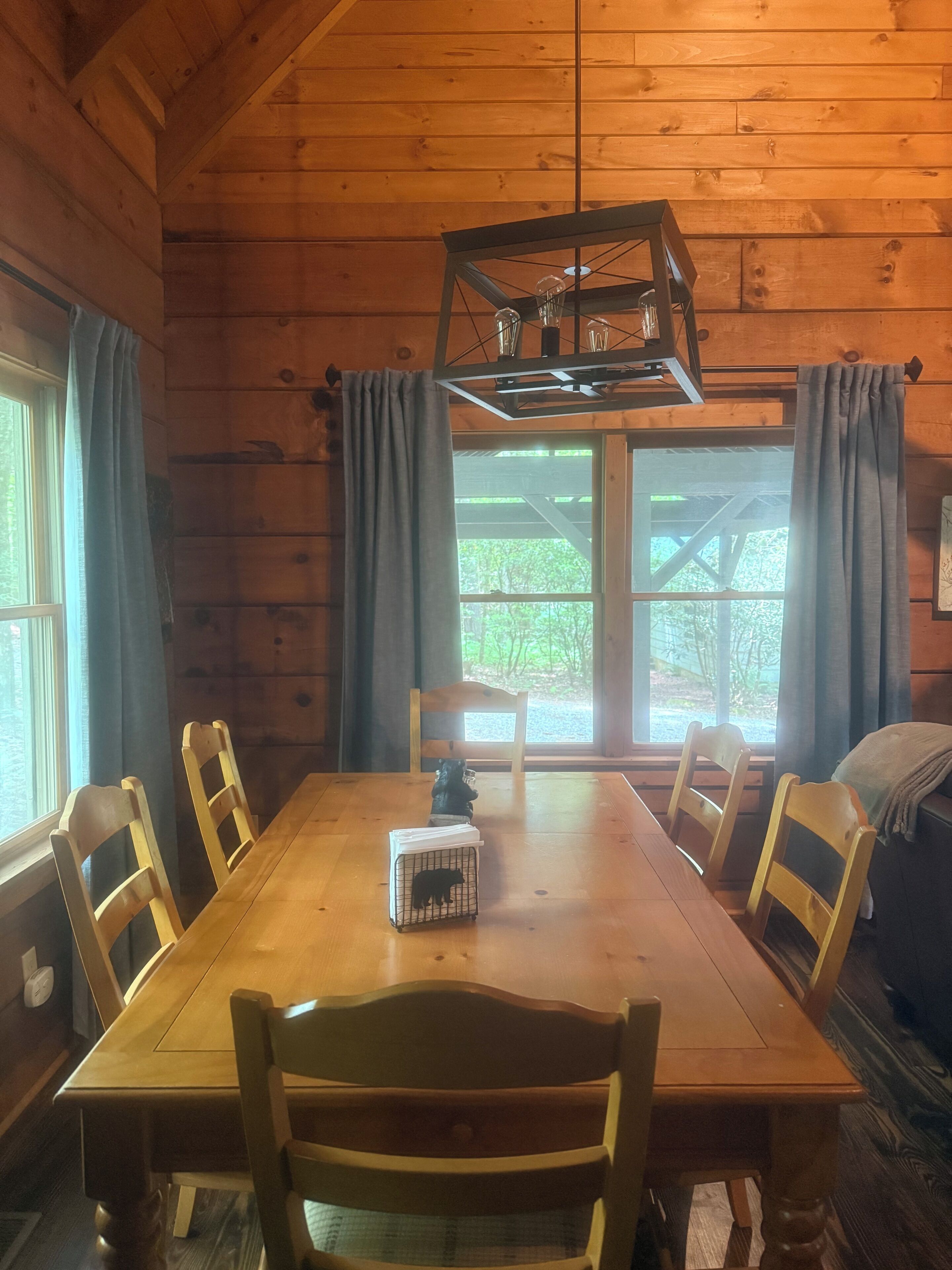 A wooden cabin interior with a dining table, chairs, a hanging light fixture, and a view of greenery outside.