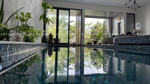 A modern indoor swimming pool with clear glass walls, surrounded by potted plants and a tiled wall.