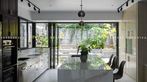 A modern kitchen with a marble island, black chairs, and a view of a garden through large windows.