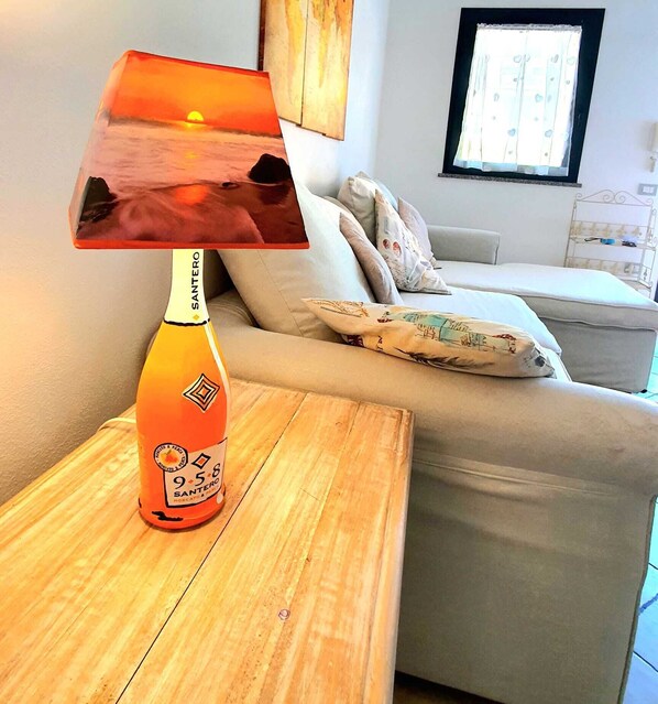 A room with a wooden nightstand, a table lamp with a sunset image, a beige sofa, and a window with blinds.
