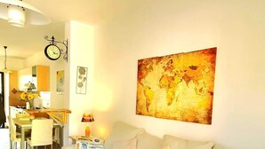 A living room with a beige sofa, a world map artwork, a clock, and a dining area with a table and chairs.