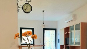 A dining area with a table, chairs, a vase with flowers, a clock, and a kitchen area with cabinets and a TV.