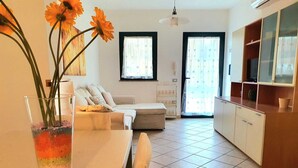 A living room with a sofa, a dining table with a vase of flowers, and a kitchen area with cabinets and a counter.