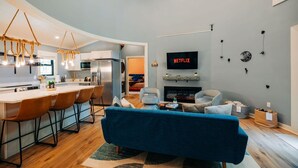 Cozinha moderna com bancada e assentos, sala de estar com sofá azul, lareira e televisão na parede.